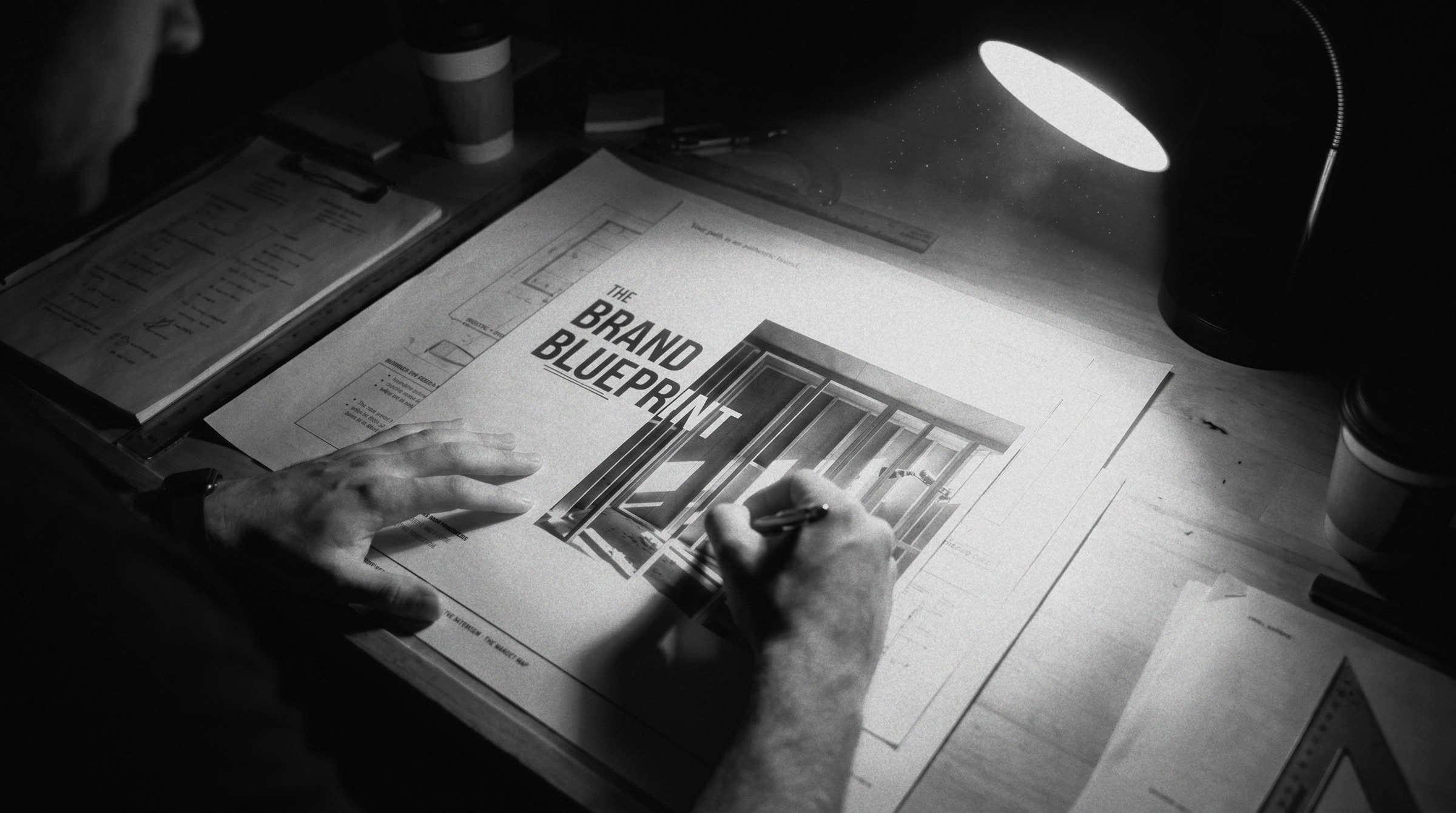 A hand drafting The Brand Blueprint document at a desk under a single lamp, annotated plans spread around it