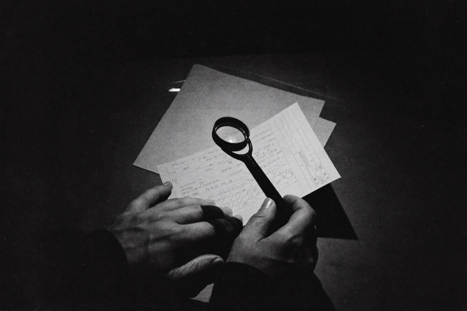 Hands examining a stack of receipts and printed charts through a loupe on a dark desk